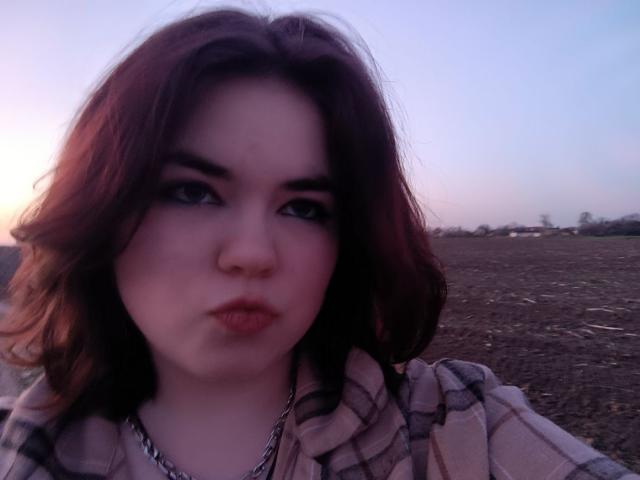AliceShoft with pouty lips, wearing a plaid shirt and chain necklace, has medium brown wavy hair, and a pale complexion, looking directly at the camera with slightly raised eyebrows.
