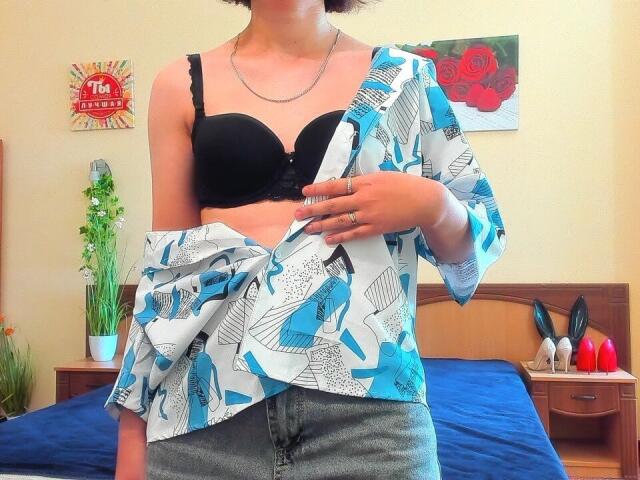Steisii with black bra, showing tits, wearing blue and white patterned shirt, gray jeans, and silver necklace, holding shirt, short curly hair, in bright bedroom.