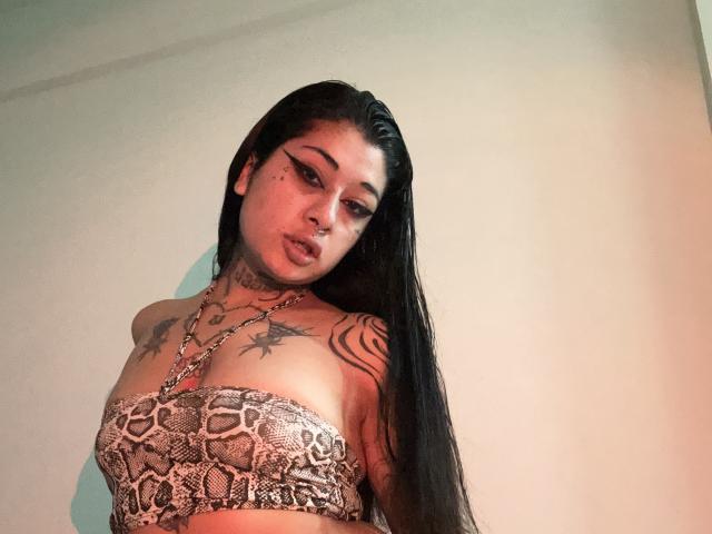 TattooMiax mit langen, schwarzen Haaren, einem BH mit Schlangenmuster, Tattoos auf Brust und Arm, starkem Eyeliner, die nach unten schaut, ernster Gesichtsausdruck, helle Haut, rotes Licht..