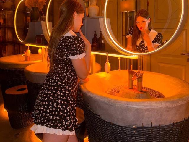 Bubochka with long brown hair, black dress with white flowers, and white trim, stands by a circular mirror, touching her chin, reflecting her red-lipped face, near a wicker sink.