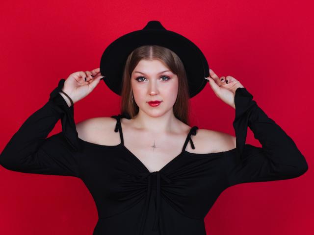 StellaLane with pale skin, blue eyes, and red lipstick, wearing a black off-shoulder dress and hat, holding it, showing small tits, and a cross necklace against a red background.