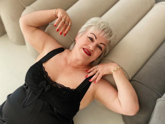KayleSmoll with short gray hair, red lipstick, black lace top, red nails, lying on beige sofa, smiling, arms raised, wearing gold bracelet and rings. Boobs visible, top open.