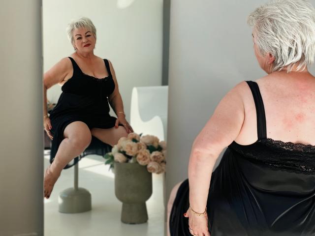 KayleSmoll, short white-haired woman, sits in black dress, showing her bare ass in mirror reflection, looking confident, with a bouquet of roses on a table behind her.