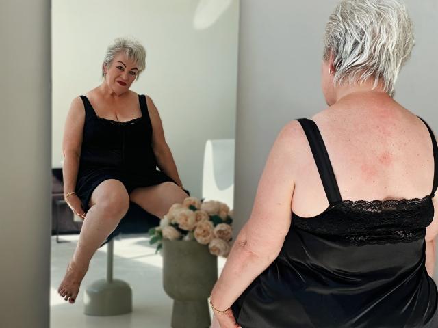 KayleSmoll with short white hair, black lace-trimmed dress, sitting on chair, reflected in mirror, smiling, showing back and bare ass, with a vase of roses in front.