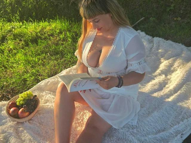 MirayDesire, con il seno prosperoso, seduta su una soffice coperta bianca, indossa un vestito bianco, sta leggendo un libro, la luce del sole la illumina, ha i capelli biondi, accanto a lei c'è una ciotola con uva e mele..