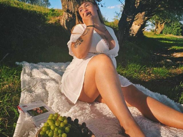 MirayDesire sitting on a white blanket, legs crossed, wearing a white dress, hands near face, grapes and a magazine in front, sunlit, tree in background, tattoo on arm.