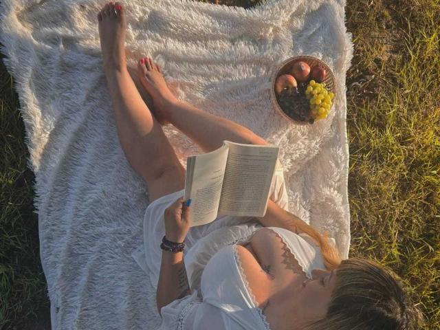 MirayDesire, sdraiata su una coperta bianca e soffice, sta leggendo un libro, indossa un abito bianco con una spalla scoperta, con i glutei e i piedi scoperti, e accanto a lei c'è un cesto di mele e uva..