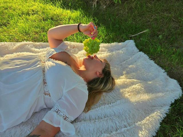 MirayDesire lying on a white fuzzy blanket, eating green grapes, wearing a white dress with floral embroidery, sunlight on her blonde hair, grass in the background.