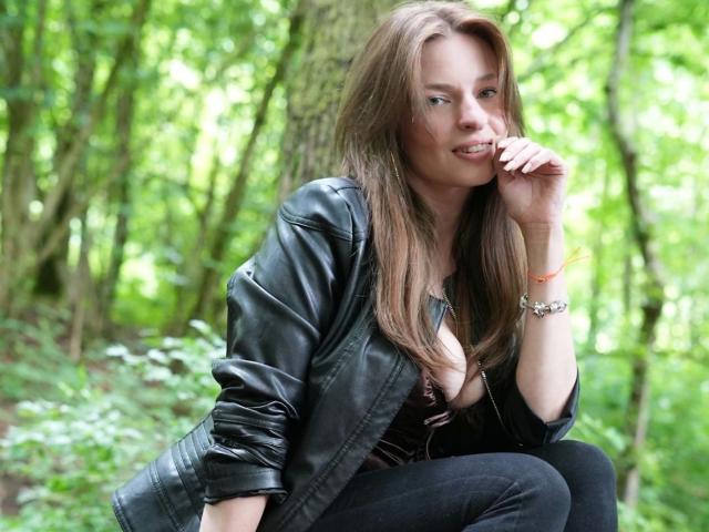KatherinePierce-hot, with long brown hair, black leather jacket, and black jeans, squats in a forest, touching her chin, smiling, showing her small tits and cute ass.