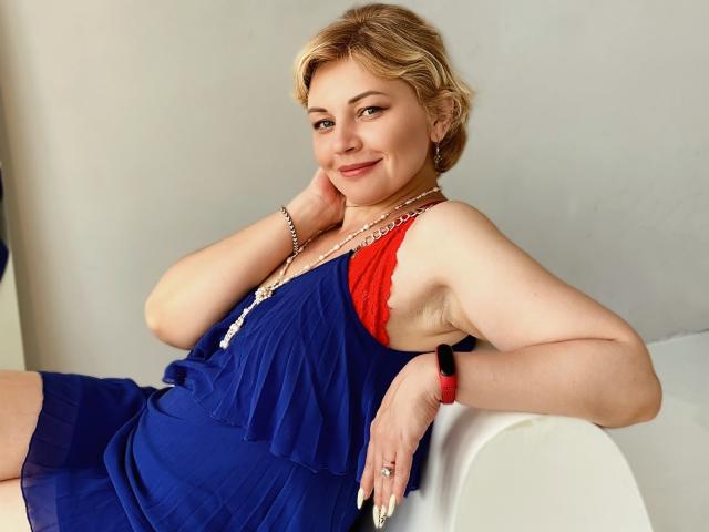 LadyElina with short blonde hair, blue dress, red accent, smiling, resting arm on white chair, wearing bracelet and red watch, fair skin, slight belly, confident, sexy pose.