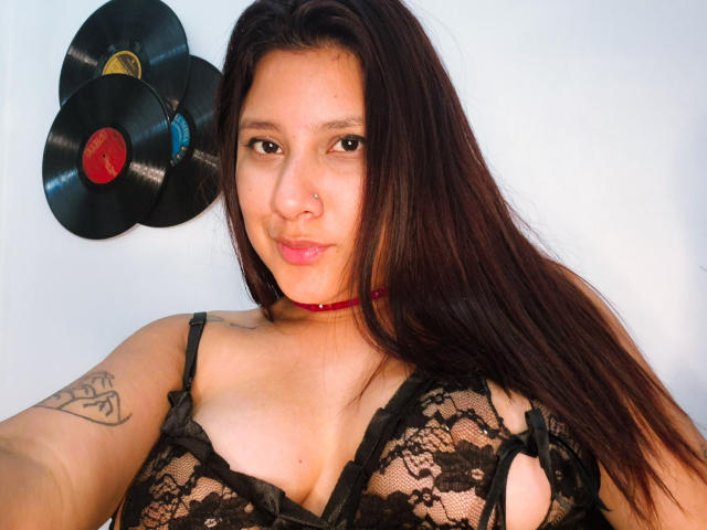 LadyStarOne, avec de longs cheveux bruns, un anneau au nez, un collier rouge, un soutien-gorge en dentelle noire laissant entrevoir ses seins, un tatouage sur le bras droit, regardant la caméra, deux disques vinyles sur un mur blanc..