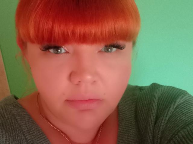 RedBustyX with red-orange bangs, green eyes, long black eyelashes, pale skin, small nose, subtle pink lips, gray ribbed shirt, gold chain, looking slightly to the side.