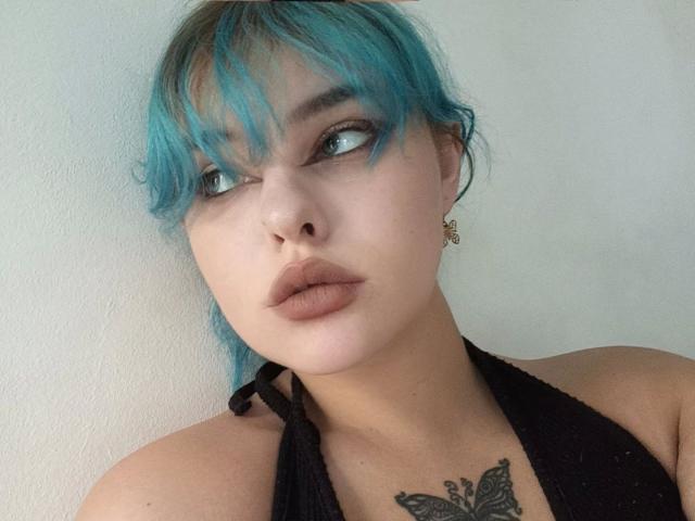 LunaGeil, met blauw haar, volle lippen en een zwarte haltertop, met een vlindertatoeage op de borst, kijkt opzij, draagt kleine oorbellen, voor een effen witte muur..