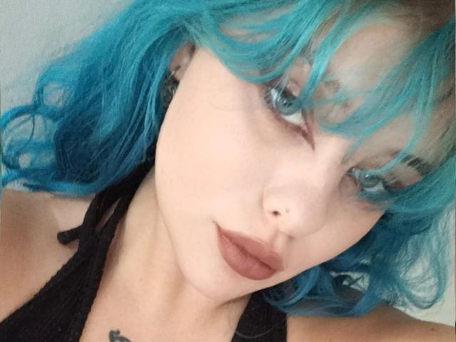 LunaGeil, aantrekkelijk met blauw haar, bleke huid en zwarte lippen, kijkt omhoog met blauwe ogen, draagt een zwarte top en heeft een kleine tatoeage onder haar sleutelbeen..