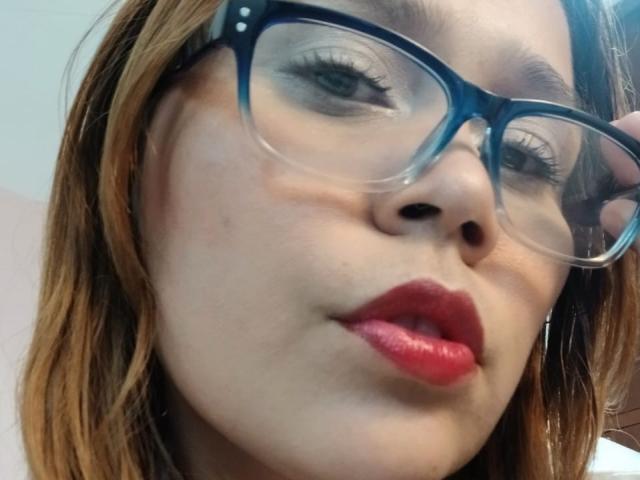 SophiaQueens has light brown skin, red lipstick, blue glasses, and brown hair in a close-up selfie. Her slightly puffed lips are centered, and she's looking down slightly.