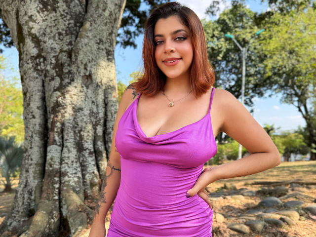 MedeaHelios with brown hair, purple dress, large tits, tattoo on right arm, standing in front of a big tree, hand on hip, smiling, sunlight, blue sky.