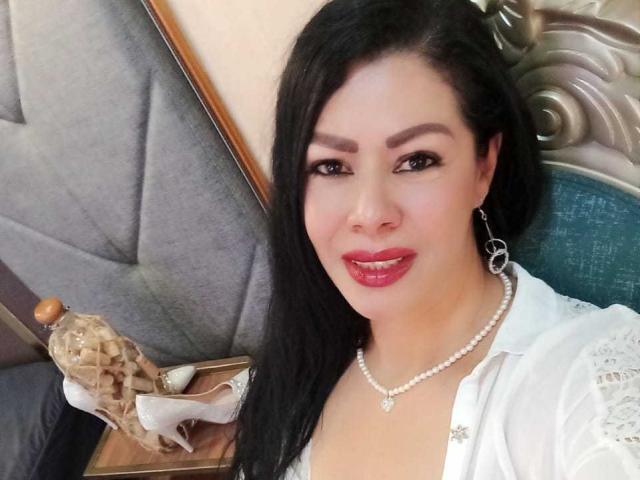 SofiMontes with shiny red lips, black hair, pearl necklace, white blouse, and silver earrings, sitting next to a beige shell and brown book. Her tits are slightly visible.