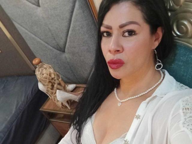 SofiMontes with long black hair, red lipstick, pearl necklace, white lace top, showing her boobs, standing next to a wooden table with a wicker basket and white heels.