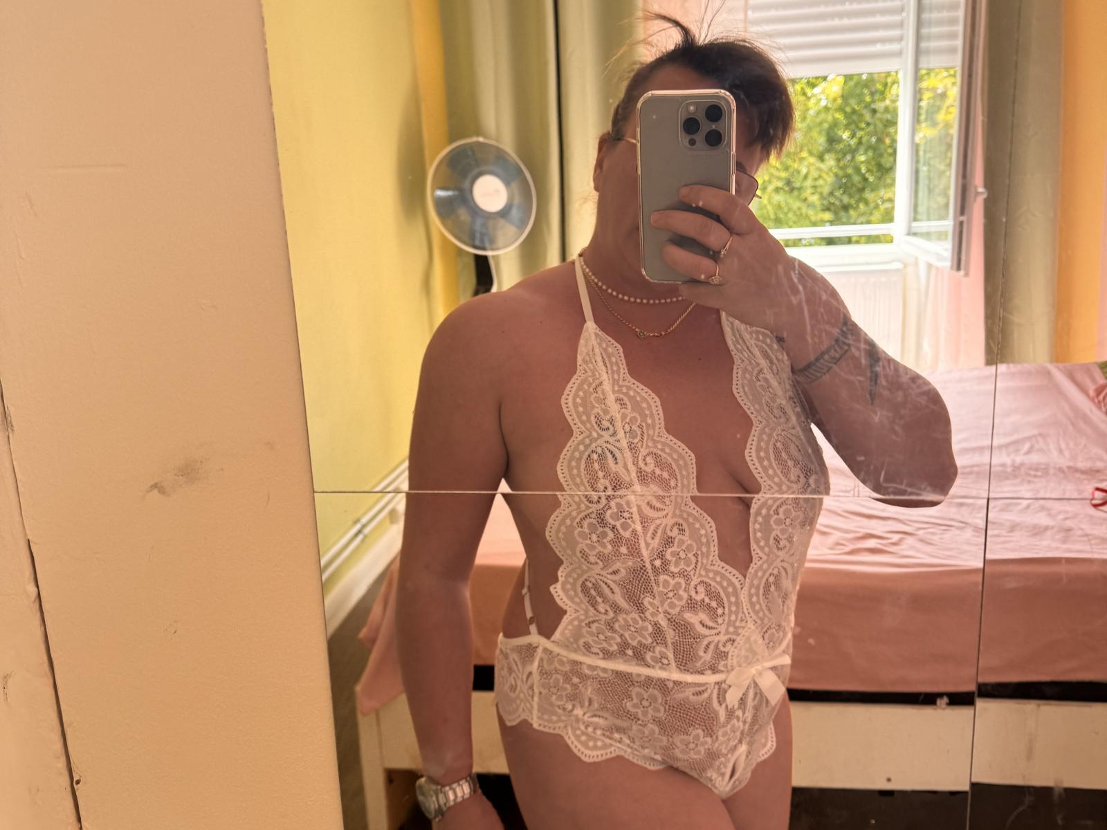 SexyRusse wearing a white lace bodysuit with a deep cleavage, showing their lace-covered pussy and small, perky tits, taking a mirror selfie in a sunlit room.