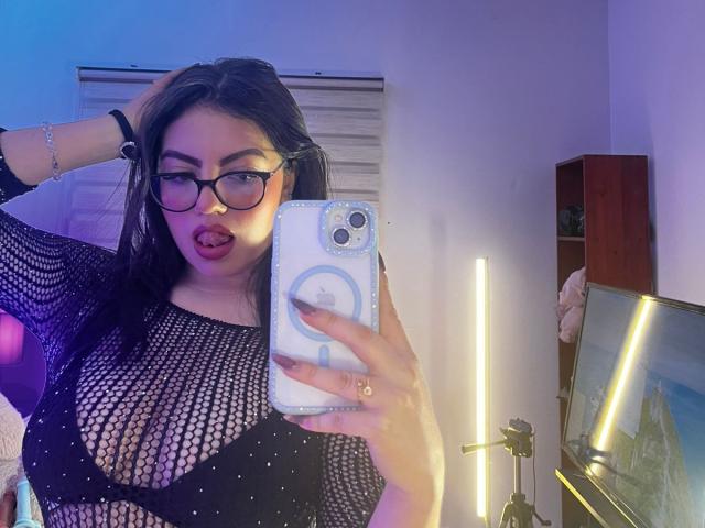DorothyLane with glasses, black mesh top showing her tits, tongue out, holding phone, wearing bracelet, in a room with purple and blue lighting, mirror selfie.
