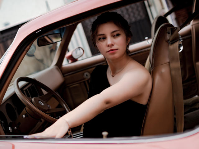 LinaFoxyHot with her ass exposed, sitting in a pink car, leaning on the steering wheel, wearing a black top, looking serious, brown hair, light skin, necklace, and bracelet.
