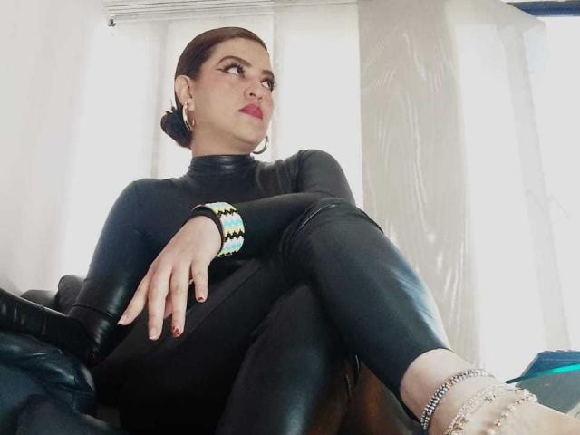 SkylarDesire with dark skin, red lipstick, and black latex outfit, sitting with legs up, showing her bare feet with red nails and silver anklet, looking to the side.