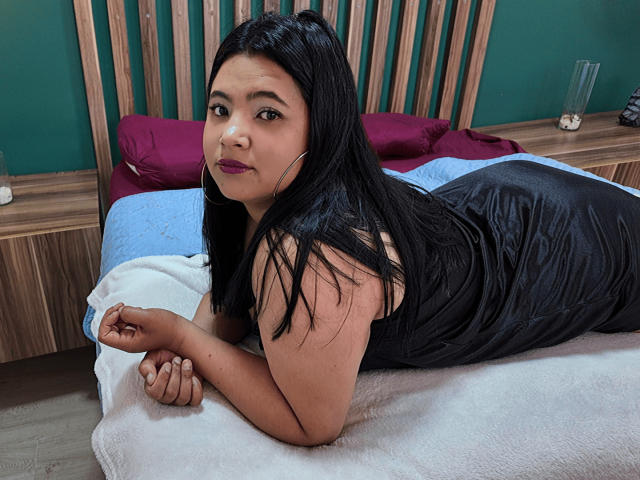 EmilyXGrace with dark hair, wearing a black lace dress, sitting on a bed, hands on thighs, showing her ass, looking at the camera, in a green-walled room.