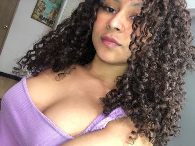 SofiaPassionne with curly brown hair, pink eye makeup, and purple lace top, showing off her boobs with a hand near her mouth, and a cute, confident smile.