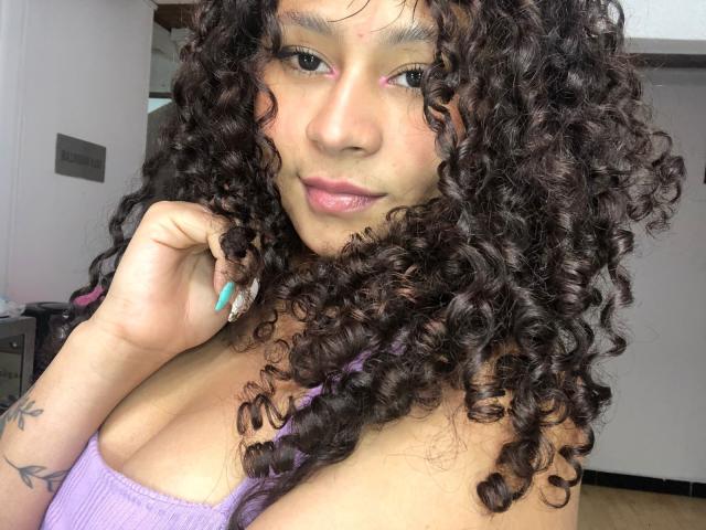Sofia, avec ses cheveux noirs bouclés, son fard à paupières rose et son haut violet, montrant ses seins, ses ongles bleus et ses lèvres roses, tenant ses cheveux près de son visage..