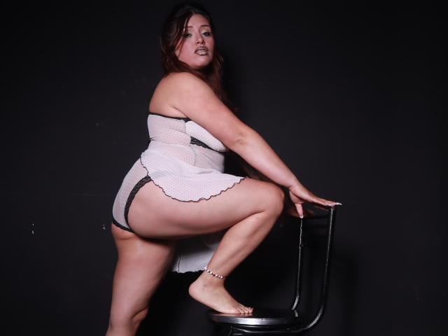 TaylorBecker with long brown hair, looking down, holding her large, bare tits with both hands, dark background, light makeup, slightly parted pink lips, and small earrings.