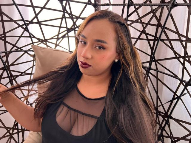 Selena Rojas, con cabello castaño largo, vestida con una blusa negra transparente que deja ver sus pequeños senos, lápiz labial rojo oscuro y una expresión relajada, sentada en una silla de mimbre negra..