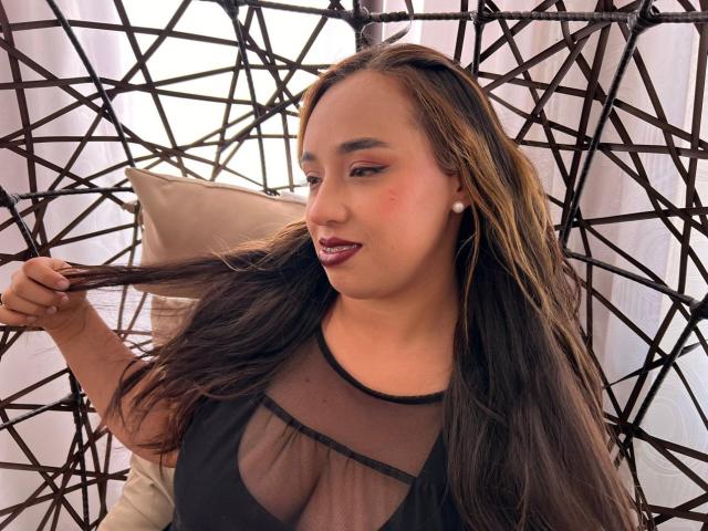 Selena Rojas, avec de longs cheveux bruns, portant un haut noir transparent, tenant ses cheveux, dévoilant sa poitrine, avec un rouge à lèvres foncé et des boucles d'oreilles de perles, se tient devant une chaise en osier..