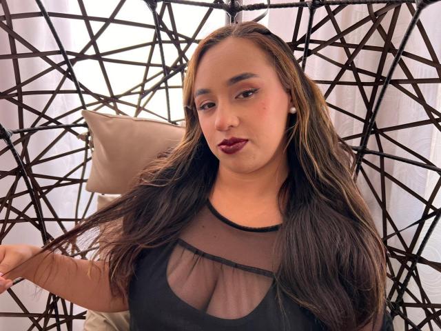 Selena Rojas, con lunghi capelli castani, rossetto scuro e una maglia nera trasparente, seduta su una sedia in vimini, che mostra il seno, con un cuscino beige dietro di lei..