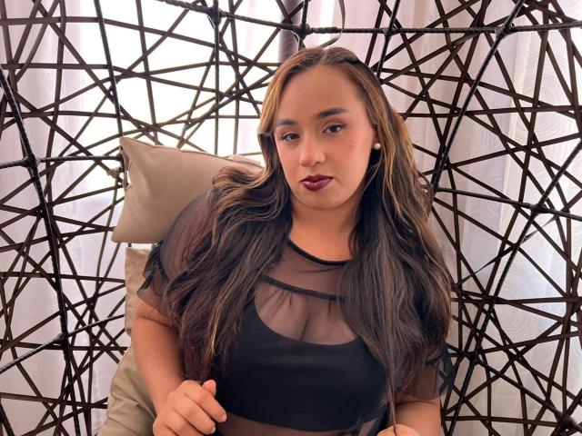 Selena Rojas, mit hellbrauner Haut, langen braunen Haaren, dunklem Lippenstift, einem schwarzen, transparenten Oberteil und Perlenohrringen, sitzt auf einem Stuhl mit einem geometrischen schwarzen Muster. Die Brüste sind unter dem Oberteil sichtbar..