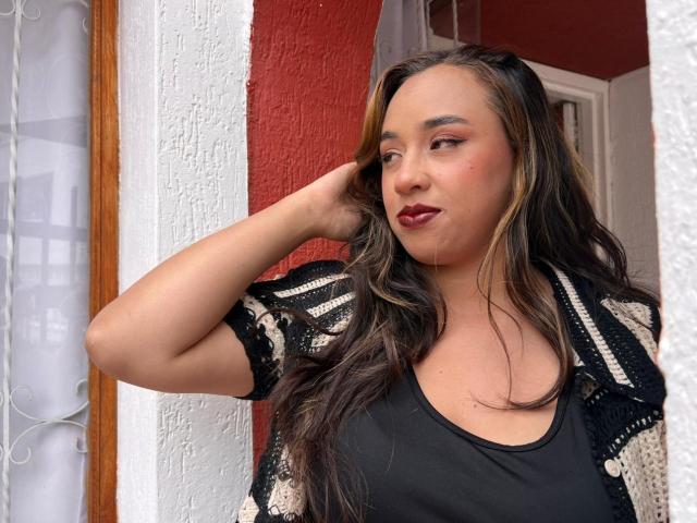 SelenaRojas with long brown hair, dark lipstick, black top, and black crochet cardigan, hand in hair, standing by white and red wall, looking to the side, confident.