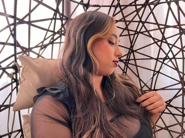 Selena Rojas, com cabelos castanhos longos, blusa preta transparente e batom rosa, olhando para o lado, com a mão no peito, cabelos ondulados, olhos castanhos, pele bronzeada, cadeira de arame estilo boho, almofada atrás..