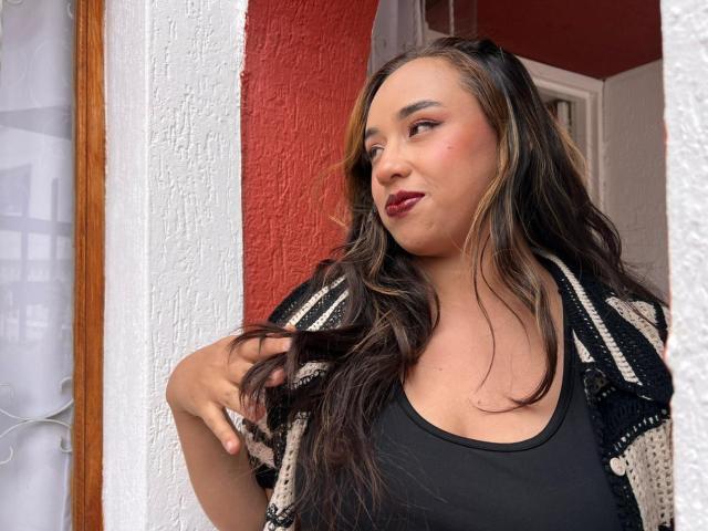 Selena Rojas ha lunghi capelli castano ondulati, indossa una maglietta nera e un cardigan lavorato a maglia nero e bianco, è in piedi con una mano sul fianco, guarda di lato e ha il rossetto rosso..
