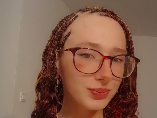 LyndaSun with braided red and brown hair, tortoiseshell glasses, and red lipstick, smiling slightly, showing a small nose ring, fair skin, and a cute, confident face.