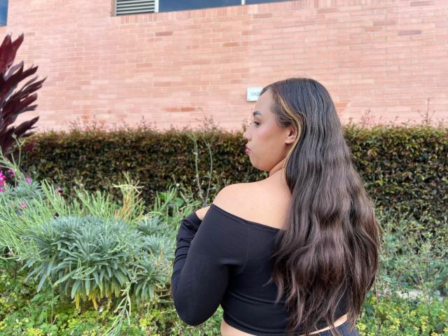 SelenaRojas with long wavy brown hair, off-shoulder black top, facing sideways, showing her back and ass, standing in a garden with green plants and a brick wall.