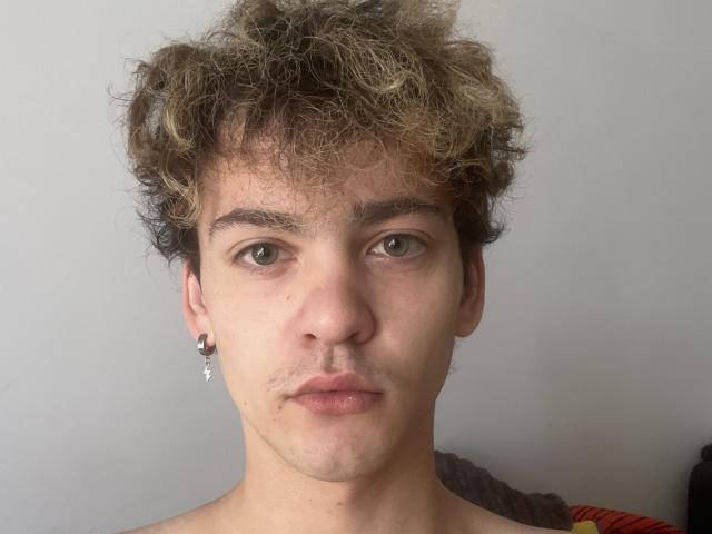 RennyCarline has curly brown hair with blonde highlights, green eyes, and a small black ear ring. Their face is serious with slightly parted pinkish lips.