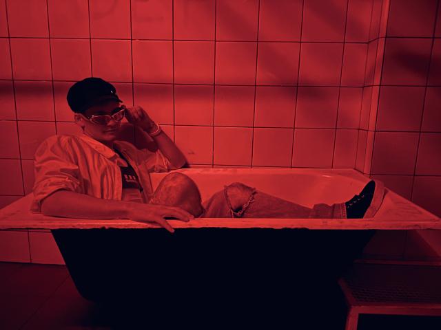 RennyCarline is in a red-lit bathroom, lounging in a black bathtub with torn jeans and black shoes, wearing a cap, open shirt, and glasses, holding a phone to ear.