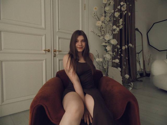 PureTalia with long brown hair, smiling, wearing a brown dress with a thigh slit, sitting on a red velvet chair, showing her bare legs and a tattoo on her arm.
