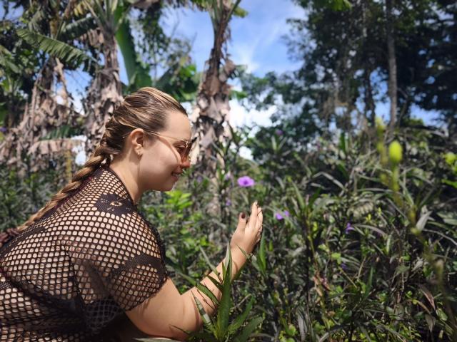 ZoeTobon69 with braided hair, wearing a black fishnet top, crouching in a lush garden, touching green plants, and smiling, with purple flowers in the background.