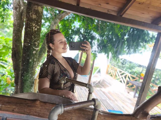 ZoeTobon69 taking a selfie, wearing a black mesh top with deep cleavage, smiling, holding a phone, standing in a wooden structure with tree in background, blonde braids.