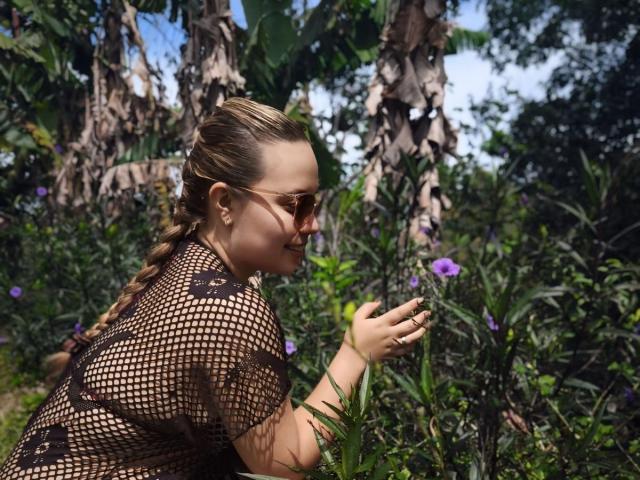 ZoeTobon69 with braided hair, wearing a mesh top, brown sunglasses, and a ring, touching a purple flower in a lush, green garden.