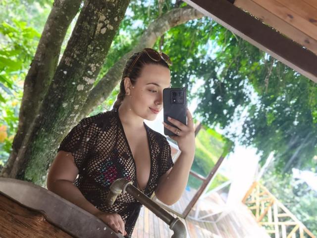 ZoeTobon69 with her tits visible through a black fishnet top, taking a selfie with a black phone, standing by a wooden counter with a faucet.