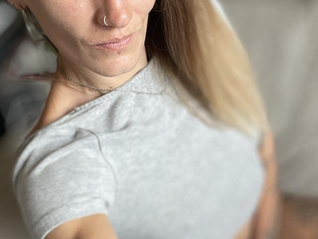 DaneeSoHot's face and upper chest, showing a nose ring, light pink lips, and a gray shirt, with blonde hair, and a silver necklace, focusing on her tits.