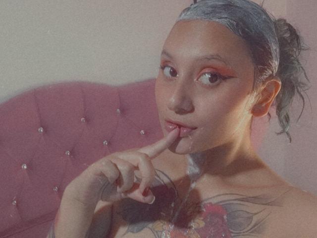 Milanixon with gray hair, red eyeliner, and finger in mouth, showing off tattoos on chest, sitting on pink tufted couch. Visible nipples, soft lighting, seductive expression.