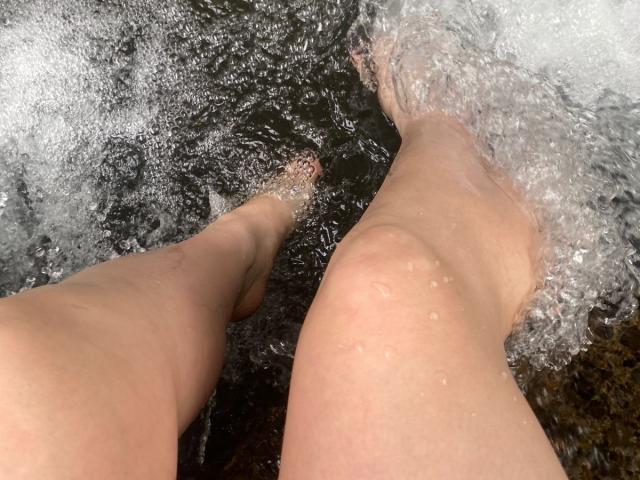 CocoVolkov's legs in water, feet partially submerged, with bubbles and splashes around. Skin is wet, showing light pinkish tone. Water is flowing, creating small waves.