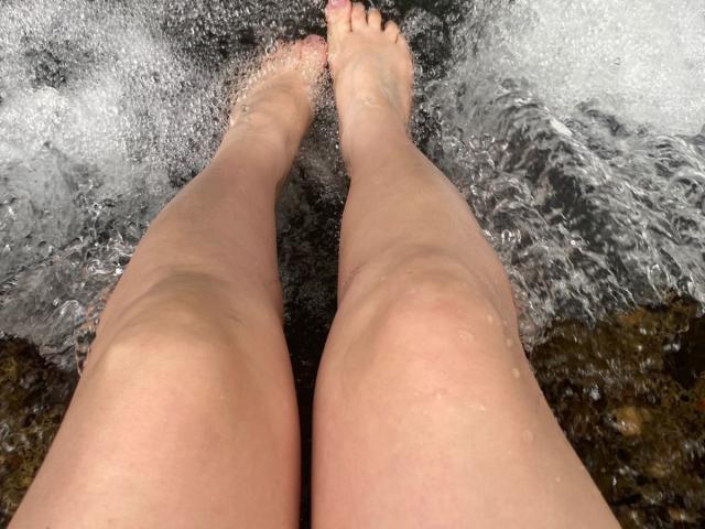 CocoVolkov's bare legs and feet standing in flowing water, with water splashing around toes. Her skin is pale and slightly wet. Her toes are pointed forward.